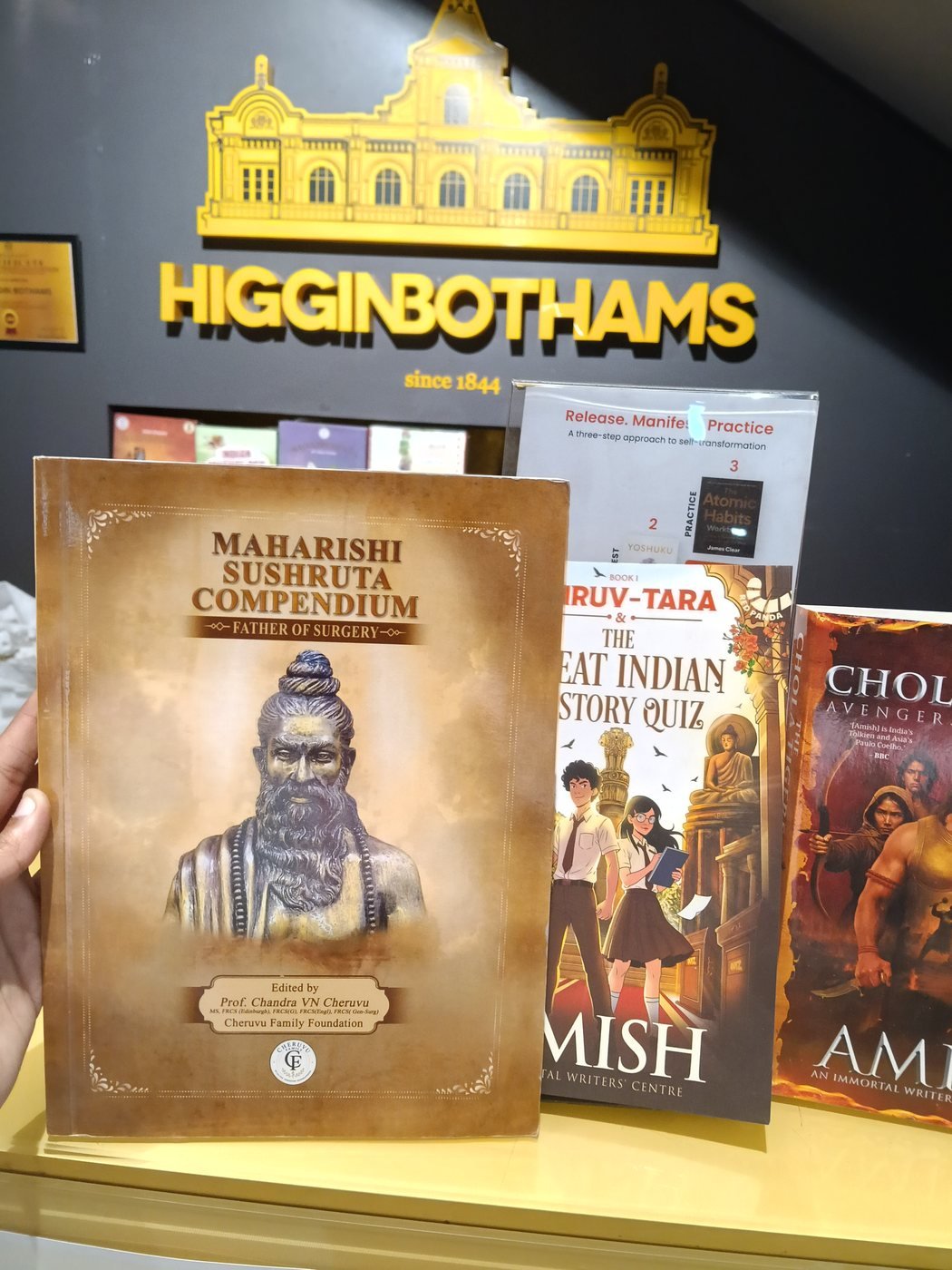 Maharishi Sushruta Compendium held up in front of the Higginbothams sign, Bengaluru